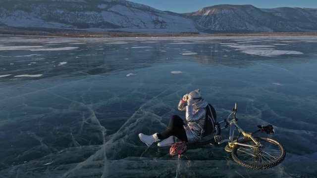 Woman is riding bicycle on the ice. The girl is dressed in a silvery down jacket, cycling backpack and helmet. The cyclist stopped to rest. She sits on the wheel and partially takes off her clothes