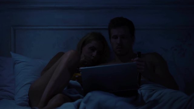 Lady and man watching horror movie on laptop, sitting on bed in dark room
