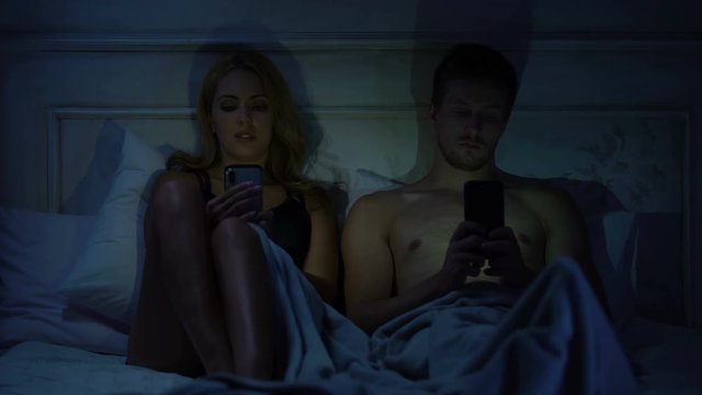 Couple sitting in bed, chatting on smartphones not talking, gadget addiction