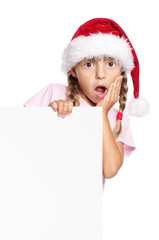 Portrait of surprised little girl in Santa hat with white blank. Child wearing Santa Claus hat, posing behind white panel. Kid holding empty Christmas billboard, peeping behind blank holidays board. 
