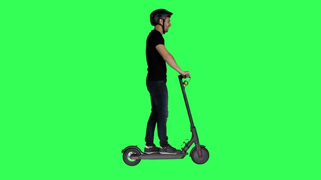 Happy young man riding an electric scooter, wearing a helmet, waves hello and looks around over a green screen.