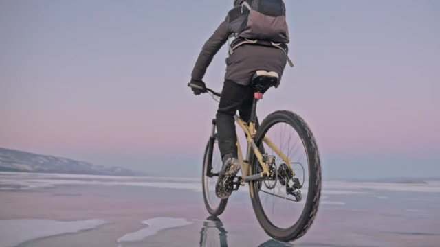 Man is riding a bicycle on ice. The cyclist is dressed in a gray down jacket, backpack and helmet. Ice of the frozen Lake Baikal. The tires on the bicycle are covered with special spikes. The traveler