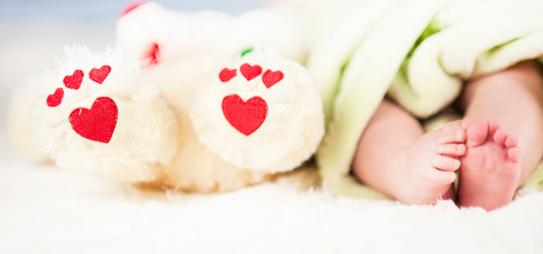 small tender legs of the baby, together with the paws of a Teddy bear with hearts on a white blanket