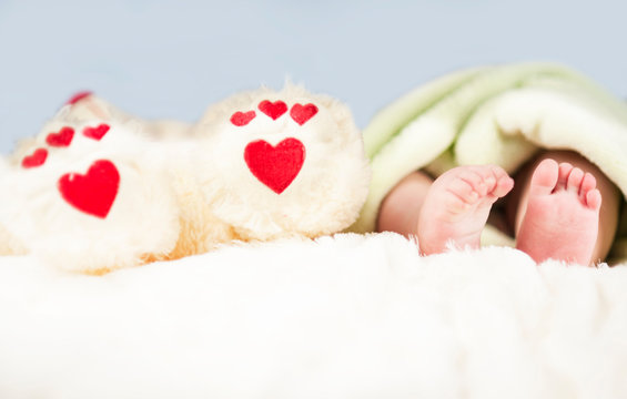 small tender legs of the baby, together with the paws of a Teddy bear with hearts on a white blanket