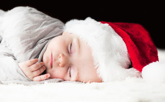 very beautiful bright happy little baby in Santa hat sleeping on white plaid wrapped in grey fabric on black background