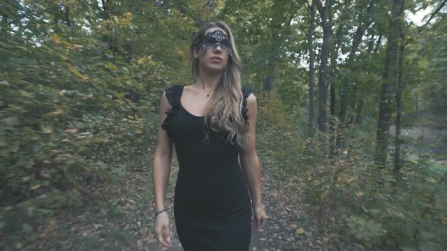 A beautiful girl in a black dress and a black mask walks along a forest autumn road.