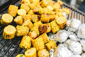 Corn cobs and potatoes wrapped in aluminum foil on a barbecue.