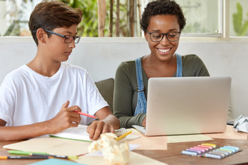 People and coworking concept. Mixed race youngsters work at school project, use digital laptop computer, write down main things and ideas in notepad, pose in desktop together. Software engineer works