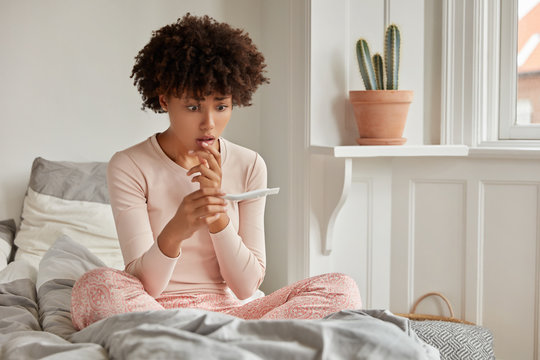 Abortion, fertility problem. Frustrated dark skinned young female holds pregnancy test which shows positive result, feels depressed and surprised, dressed in pajamas, sits at comfortable bed.