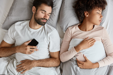 Top view of addicted mixed race family couple have deep sleep, hold modern electronic gadgets in hand, being always in touch, lie at comfortable bed, have good rest. People and technology concept