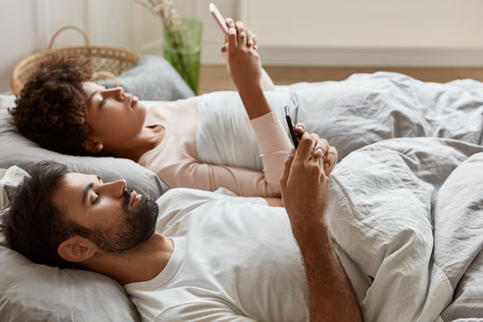 Relaxed serious couple share photos from telephone, update profile and view pictures in social networks, use high speed internet connection at home, enjoy comfort under bedclothes. Networking concept