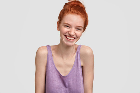 Lovely young freckled ginger female giggles at positive news, has foxy hair, shows bare shoulders, wears casual vest, stands against white studio background, expresses happiness. Studio shot