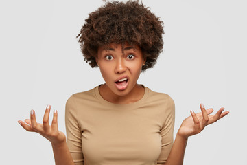 Photo of confused hesitant young woman has Afro curly hairstyle, shruggs shoulders, frowns face in...