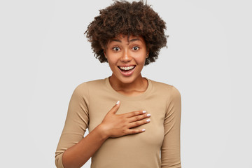 Thrilled glad dark skinned woman keeps hand on chest, surprised to recieve compliment from...