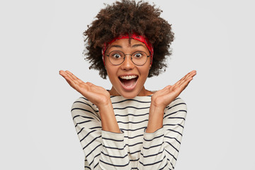 Photo of impressed amazed beautiful woman gestures with both hands, spreads hands near face, wears round glasses, striped sweater and red headband, models alone in studio, expresses surprisement