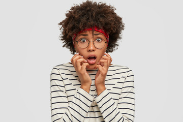 People, fright concept. Scared Afro American model gestures with anxious expression, horrified to see something horrible near, has bated breath, dressed in casual outfit, stands indoor over white wall