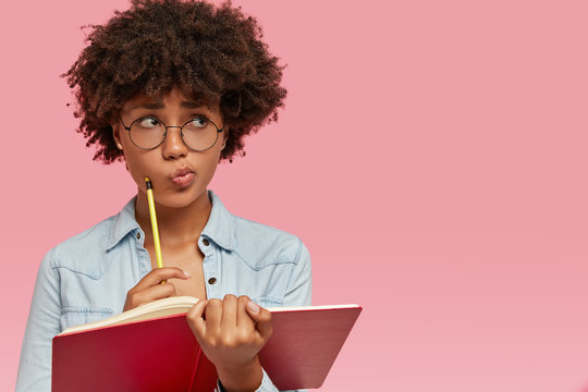 Pensive confused dark skinned woman purses lips in bewilderment, writes notes in notepad, thinks on creating new article for personal blog, wears round transparent glasses poses indoor with copy space