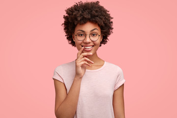 Fototapeta na wymiar Waist up shot of pleasant looking smiling Afro American lady has happy expression, keeps hands near mouth, rejoices recieving present, dressed in casual t shirt, isolated over pink background