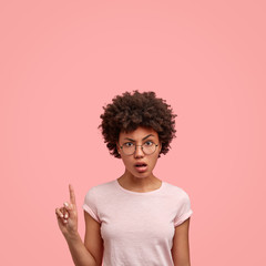 Indignant dark skinned curly lady points with index finger upwards, attracts your attention to blank copy space, dressed in casual t shirt, isolated over pink studio wall, suggests go upstairs