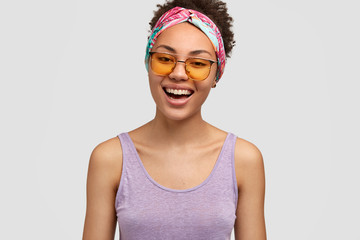 Positive dark skinned woman with cheerful expression, smiles broadly at camera, wears casual vest and trendy sunglasses being in high spirit, isolated over white background. People and good emotions