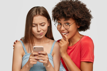 Studio shot of embarrased Caucasian woman looks at screen of mobile phone, texts messages, curious dark skinned girl embraces and looks with great interest, pose together against white background