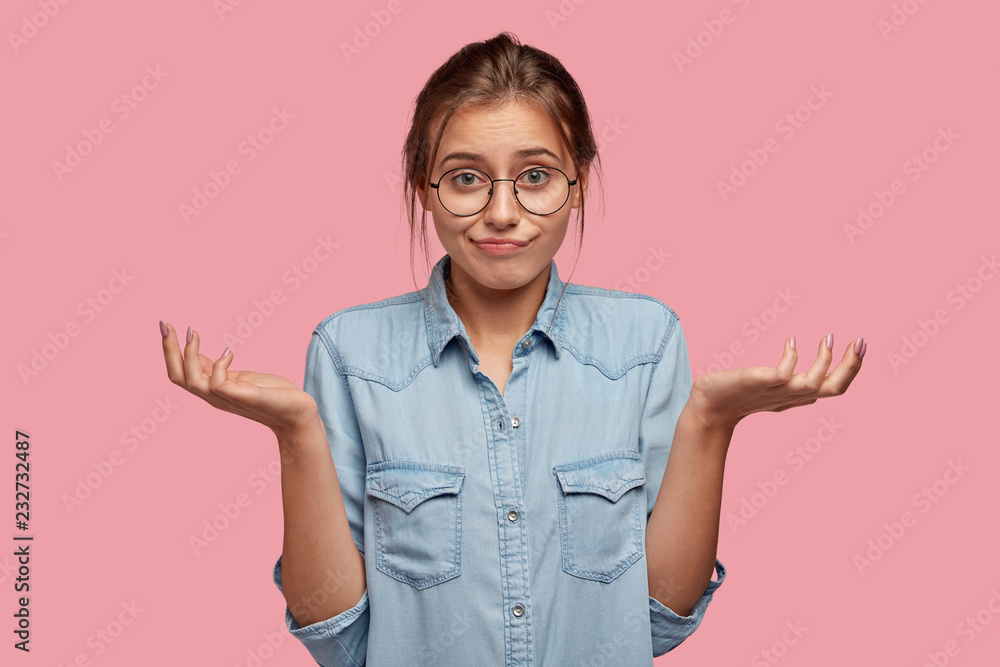 Wall mural Hesitant uncertain woman shrugs shoulders, has displeased unaware expression, cant make choice between two items, wears round spectacles, models against pink background. Who cares in this situation?