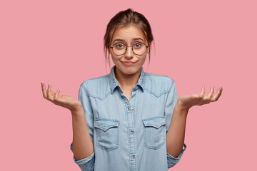 Hesitant uncertain woman shrugs shoulders, has displeased unaware expression, cant make choice between two items, wears round spectacles, models against pink background. Who cares in this situation?
