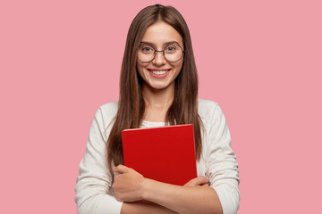 Waist up shot of pretty girl smiles pleasantly, carries red textbook closely, wears white casual jamper, models against pink background. Headshot of Caucasian lady student happy after having classes