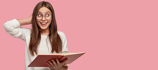 Photo of joyful attractive girl looks happily aside, wears white sweater, round eyewear, reads information from textbook, stands against pink background with free space for your advertisement