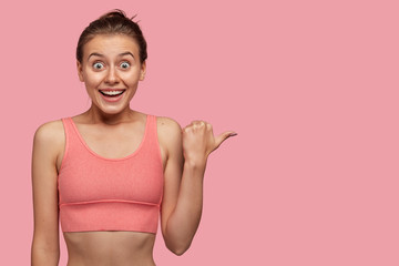 Sportswoman with amazed facial expression, being astonished by good news, indacates with thumb aside, dressed in casual top, has sporty body, isolated over pink studio background, advertises item