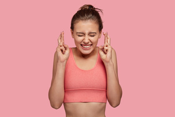 Glad wishful young woman has healthy body shape, keeps fingers crossed, eyes closed, believes in something positive, prays for good luck or wellness, dressed in pink top, models and gestures at studio