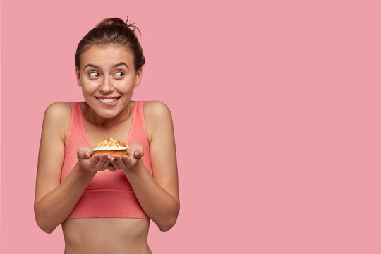Photo of attractive joyful female bites lips, holds delicious cake, feels hungry after cardio training, dressed in casual top, isolated over pink background with copy space for your advertisement