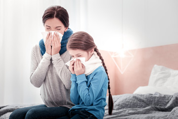 Falling ill together. Portrait of young mother and little daughter sitting on the bed while using...