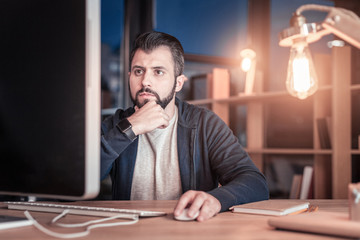 Need to think. Attentive bearded man pressing lips and looking at screen of computer while doing his job