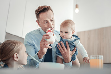 Happy father. Careful attentive occupied father holding a bottle supporting baby and playing together in the kitchen.