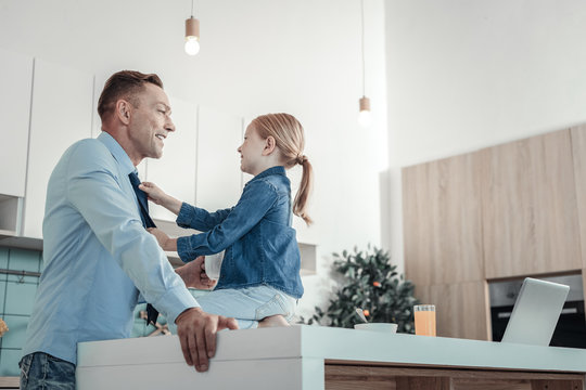 Time to go. Caring lovely pleasant father holding a cup smiling and standing in the kitchen near daughter who sitting on the table.