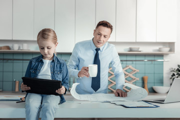 Smart generation. Serious busy pretty girl sitting in the kitchen on the table holding and using...