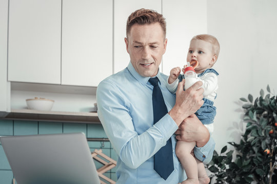 Family firstly. Occupied focused pleasant man holding a baby being at home and looking at the laptop.