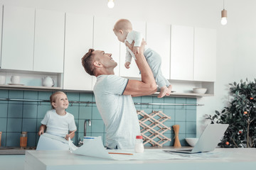 The happiest father. Joyful lovely reliable father standing in the kitchen near his daughter smiling and lifting a baby.