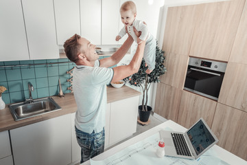 Reliable support. Satisfied strong pleasant father standing in the kitchen lifting a baby and...