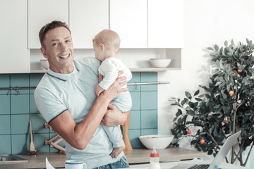 Be happy. Joyful handsome lovely father standing in the specious room smiling and holding a baby.