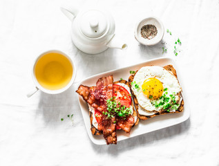 Served breakfast - toast with cream cheese, fried egg, tomatoes, bacon and green tea on a light background, top view