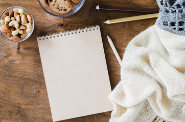Flat lay of blank brown notebook with knitted blanket and snacks on wooden table.