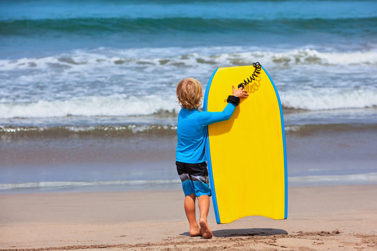 Baby boy - little surfer run with bodyboard to sea for riding on waves. Active family lifestyle, kids watersport lessons, swimming activity in adventure surfing camp. Beach summer vacation with child.