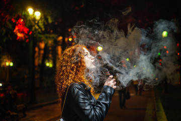 Young red-haired girl thoughtfully smokes an electronic cigarette. She walks alone on streets of night city. Non-nicotine cigarette. Wet asphalt after rain.