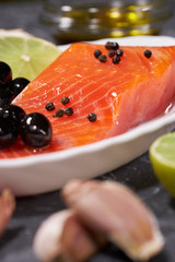 A piece of salmon on a white plate with olives and lemon, on a gray background. Selective focus