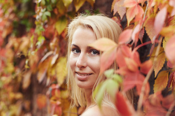 Authentic shot of European pretty blond female, mysteriously looking at camera, smiling tenderly and blissful, surrounded with autumn leaves, posing on colorful natural wall background. Beauty concept