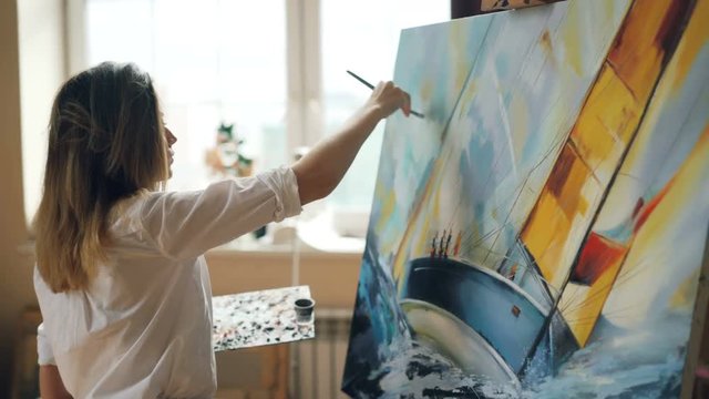 Good-looking girl artist is busy painting seascape on canvas using color palette, brush and canvas on easel working in nice studio. Talented people and work concept.
