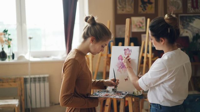 Female student of art school is painting flowers with oil paints while her teacher good-looking young woman is giving her advice pointing at picture.