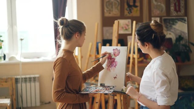 Beautiful blond girl is depicting flowers at art class under guidance of experienced teacher standing together near easel. Painting tools and artworks are visible.